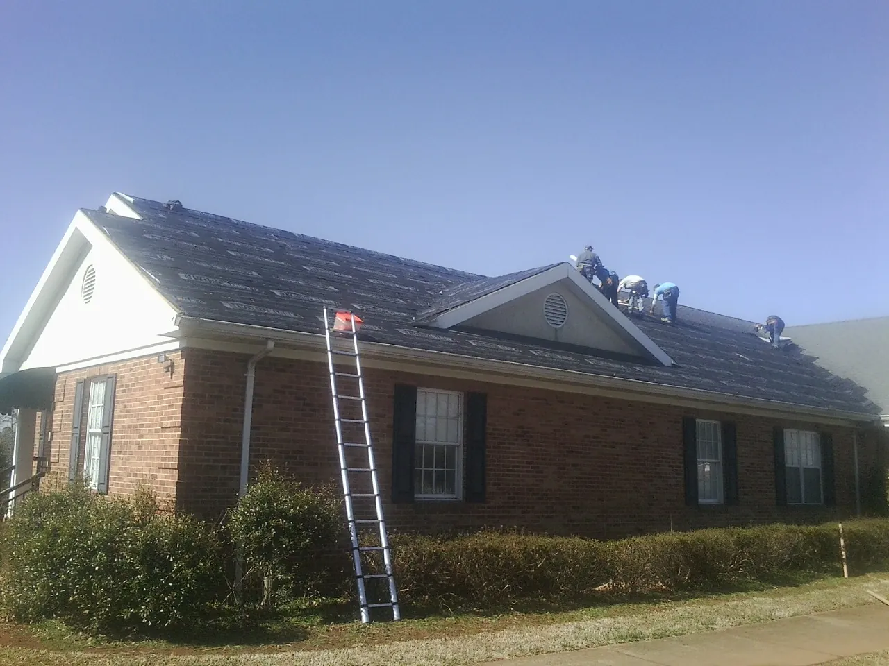 Crew tearing shingles off a brick ranch roof with a ladder set — Metal Roof Repair work by our crew in Grand Valley