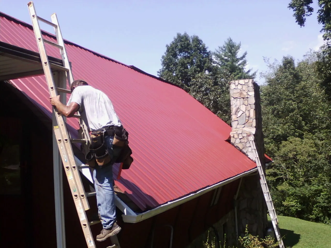 Roofer on a ladder beside a red metal roof and stone chimney — Soffit & Fascia Repair by Roofing Brothers in Grand Valley